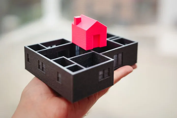 검은 3D 프린트 평면 모형 위에 빨간 집 모형을 올려 든 손