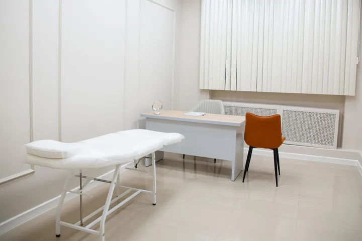 베이지 바닥 위에 진료 침대와 책상, 주황색 의자가 놓인 넓은 진료실