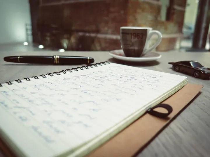 스프링 노트에 손글씨가 적혀 있고 만년필과 찻잔이 놓인 책상